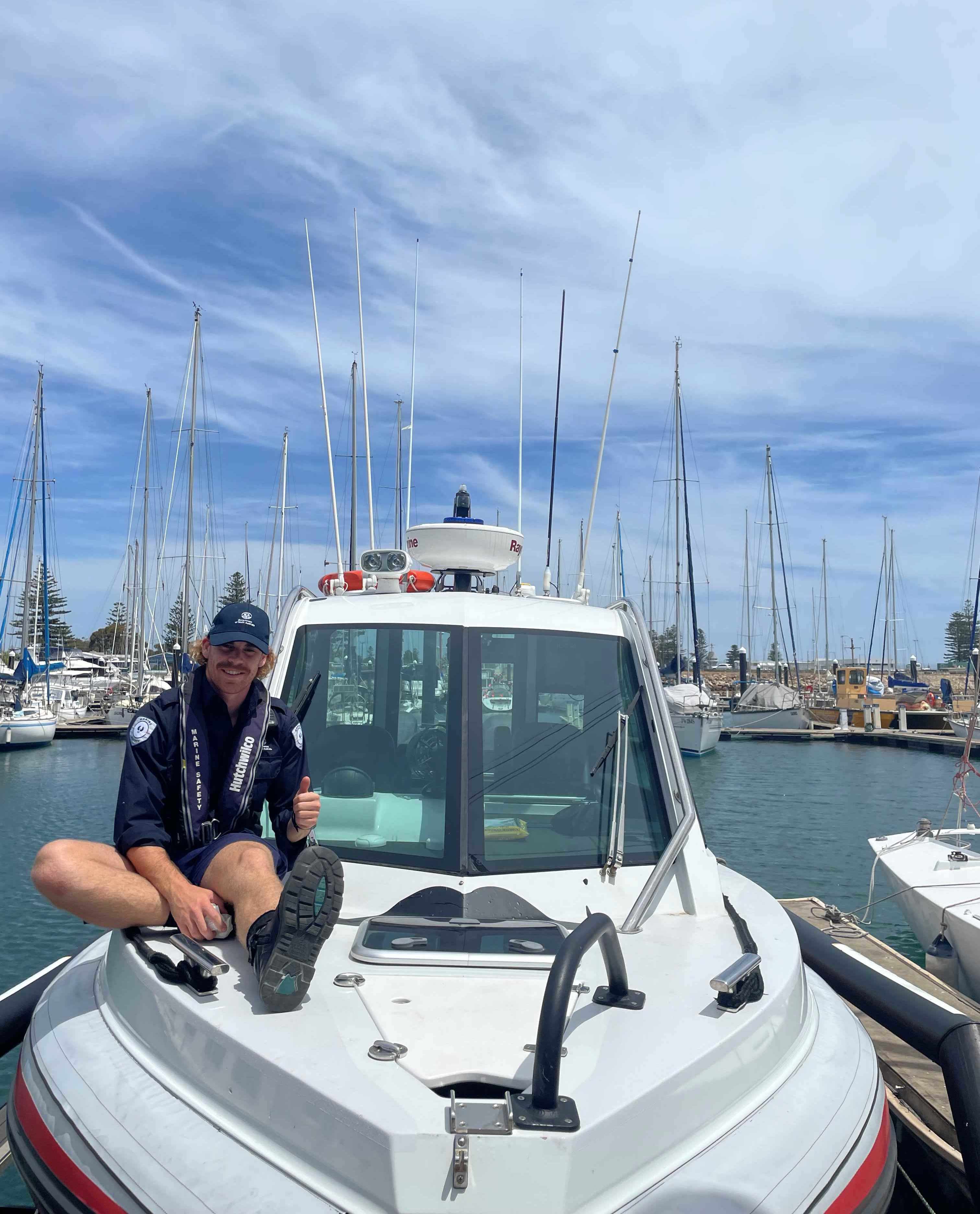 Supporting mens health. Pictured is a Marine Safety officer sitting on the front of the boat smiling with a thumbs up. Blue sky and clouds with vessels and yachts in the background.