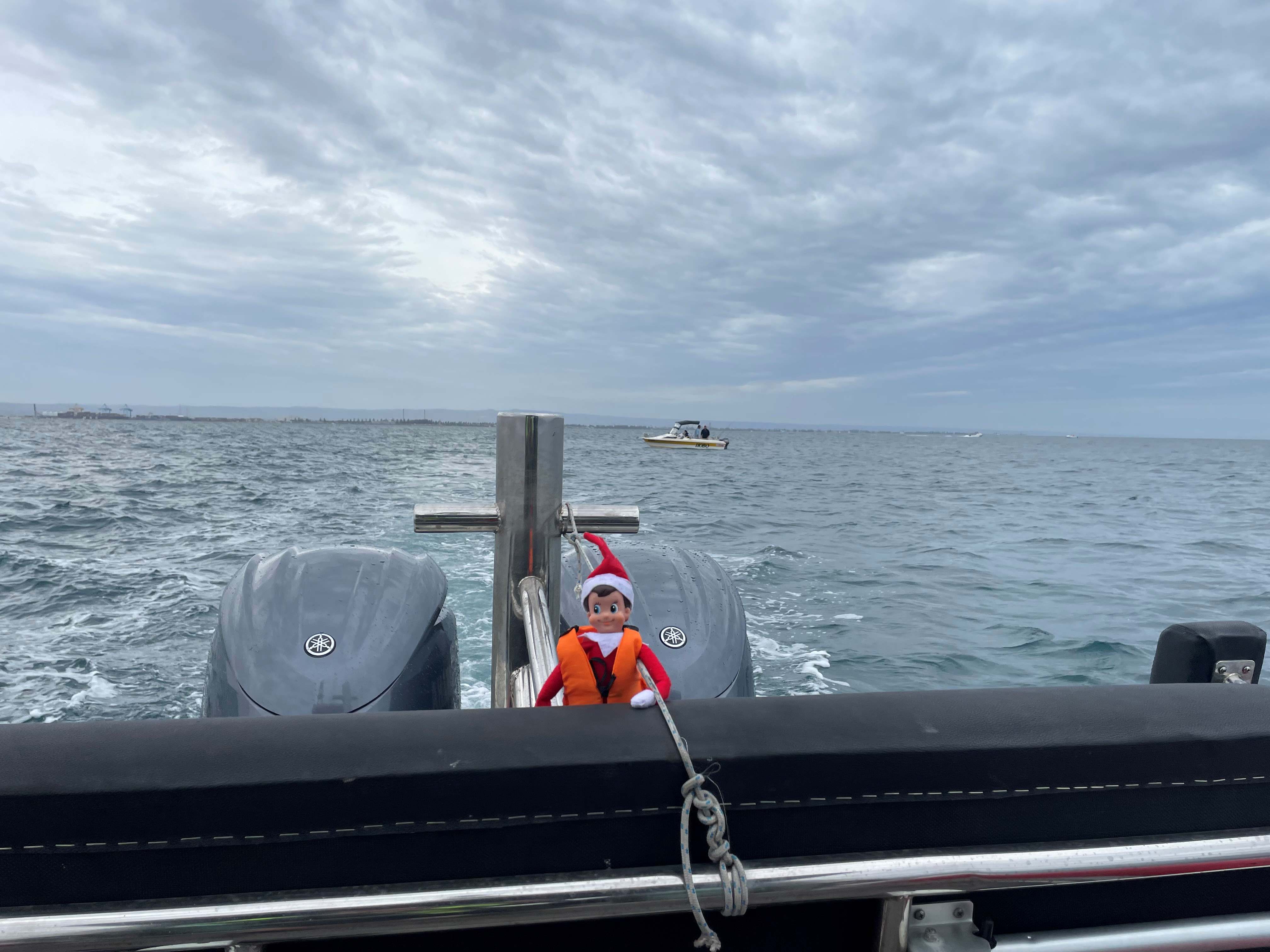 Skipper the Elf hanging onto the back of the boat with another vessel on water in the background