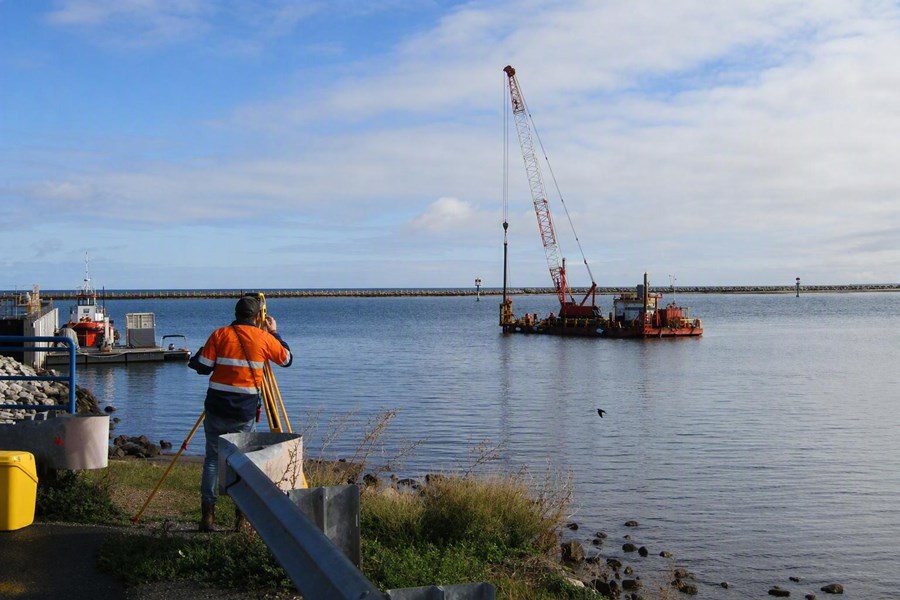 The image shows marine works being carried out at Outer Harbor, South Australia, near the entrance to the Port River and the Royal South Australian Yacht Squadron (RSAYS). In the foreground, a surveyor stands on the shoreline operating a surveying instrument mounted on a tripod. The surveyor is positioned beside a paved waterside path, with safety barriers, rocks and coastal vegetation along the shoreline.  On the water, a floating work barge supports a large crane with a tall lattice boom extending high into the sky. The crane appears to be engaged in marine construction or maintenance work within the harbour. Several navigation markers are visible in the water behind the barge, helping guide vessel traffic through the harbour entrance.  To the left of the image, a small service vessel is moored alongside a floating pontoon. In the background, a long breakwater stretches across the horizon, protecting the harbour from offshore swell. The water is calm, reflecting the overcast sky above. The photograph highlights surveying and construction activities associated with maintaining marine infrastructure at the entrance to the Port River.