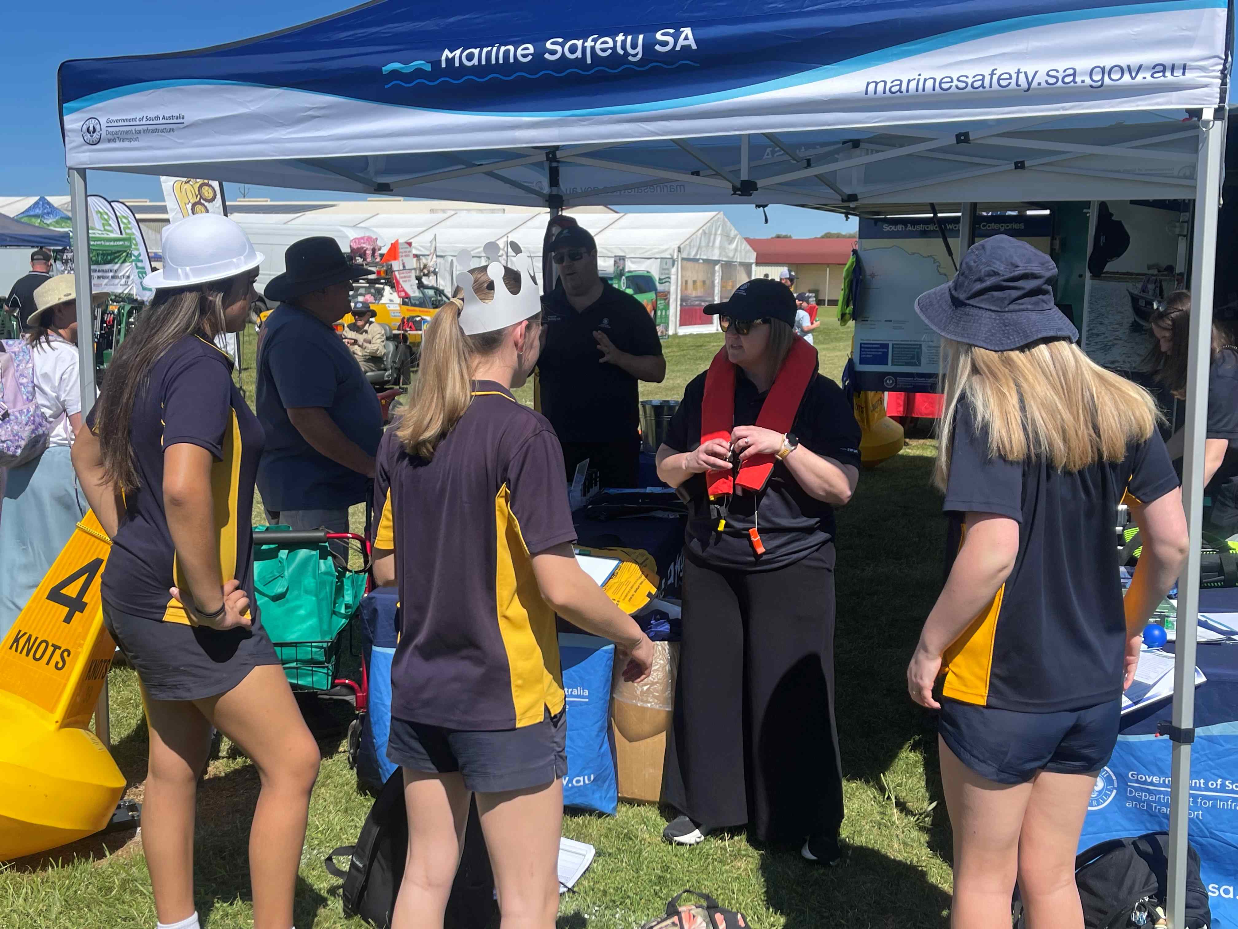 Visitors gather at a Marine Safety SA display under a branded marquee at an outdoor community event. A Marine Safety SA staff member demonstrates the correct way to wear and adjust a lifejacket while speaking with a group of attendees. The display includes boating safety information, educational resources and marine safety equipment. A yellow navigation marker buoy is visible beside the marquee, and other event stalls can be seen in the background on a sunny day.