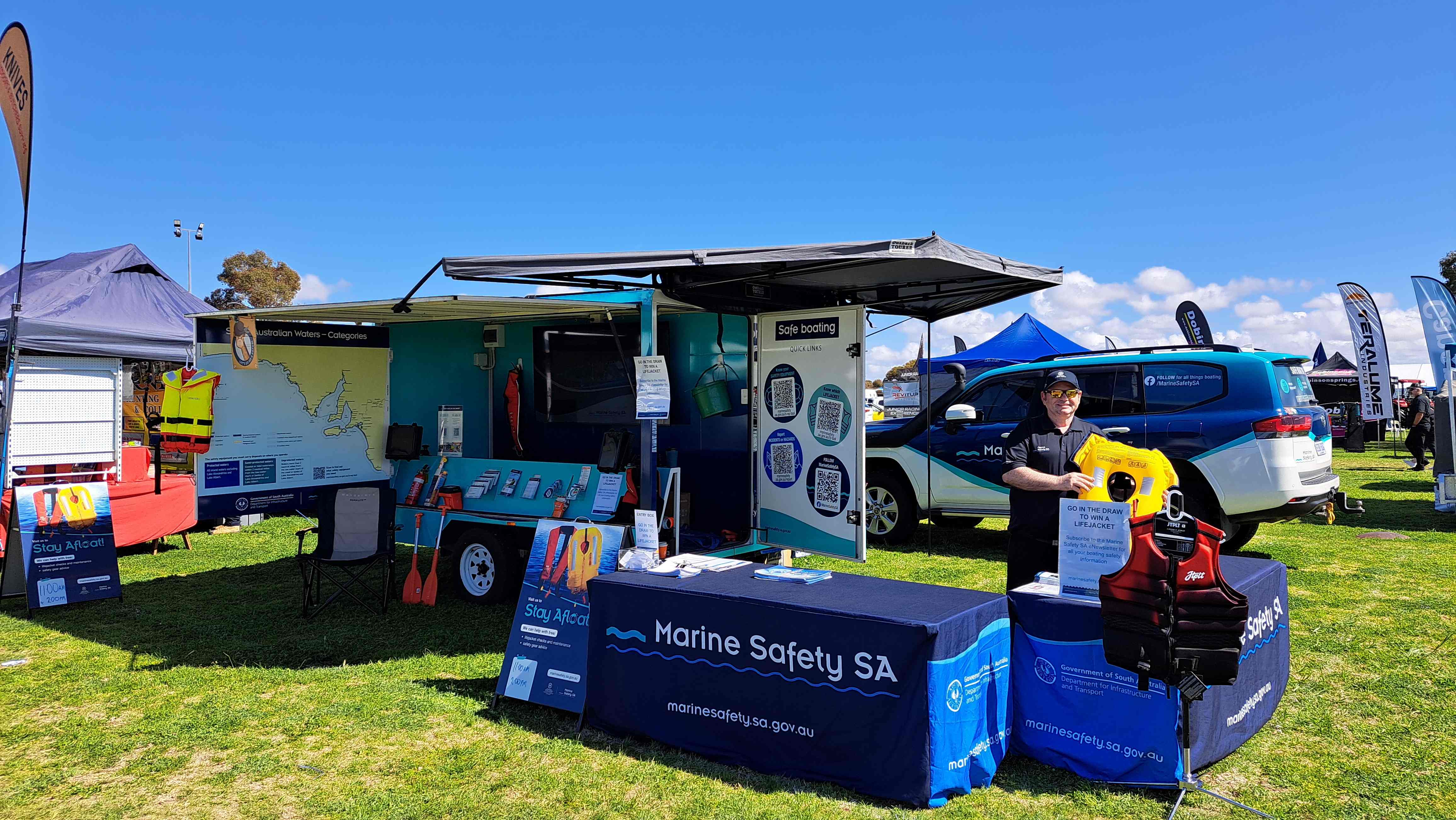 Man standing with a lifejacket at the Marine Safety SA education trailer with tables set out and the Marine Safety SA car in the background