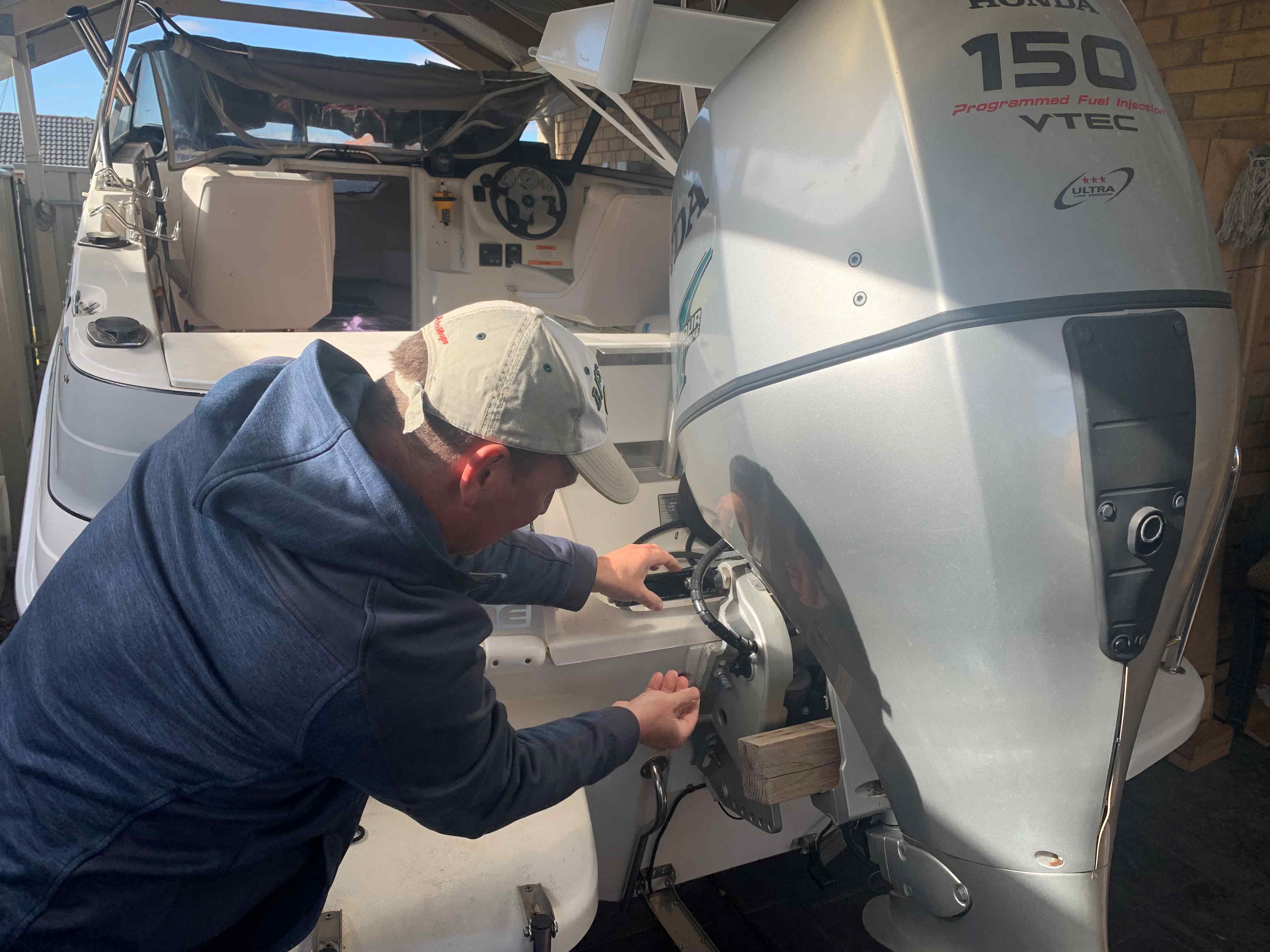 A gentleman in a blue hooded jumper and cream coloured cap checking his vessel engine and making sure everything is maintained for a safe boating trip.