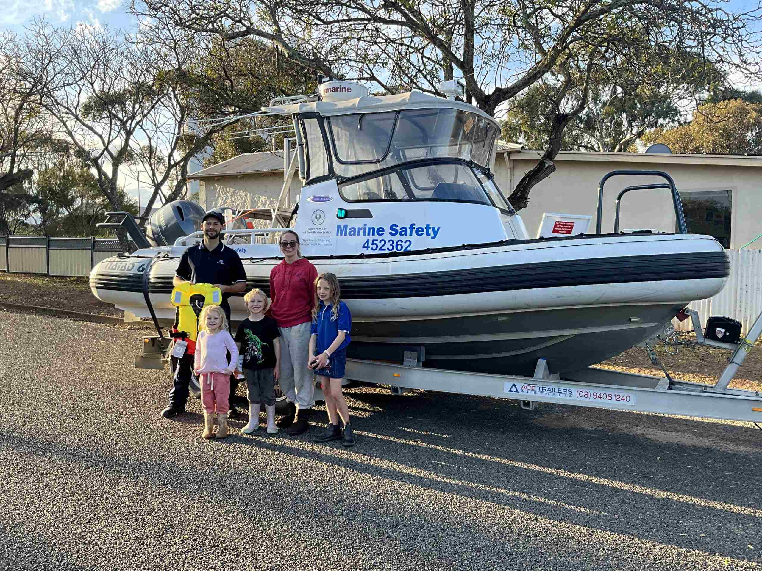 Marine Safety Officer presenting lifejacket to mother and three children in front of a Marine Safety boat on a trailer on the road