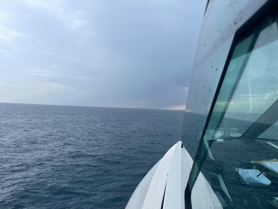 The image is taken from aboard Wardana, a Marine Safety SA vessel operating at sea. The photograph looks forward along the starboard side of the vessel, with part of the white wheelhouse and large cabin windows visible in the foreground. Water droplets can be seen on the windows, suggesting recent rain or rough weather conditions.  The vessel is travelling across open water under a dramatic sky filled with dark storm clouds. The sea is a deep blue-grey, with small waves and ripples extending to the horizon. In the distance, a faint strip of land is visible beneath the cloud cover. The image highlights the challenging conditions that boaters may encounter while conducting activities on South Australian waters.