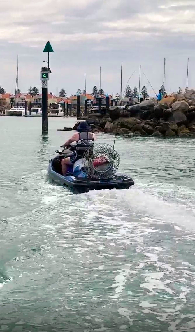 Pictured is a person on a PWC (Jetski) operating at 4 knots as the electronic sign has picked up his speed and advised he is doing 4 knots. The water has a very small wake (movement) with rocks on the side and boats moored in the background.