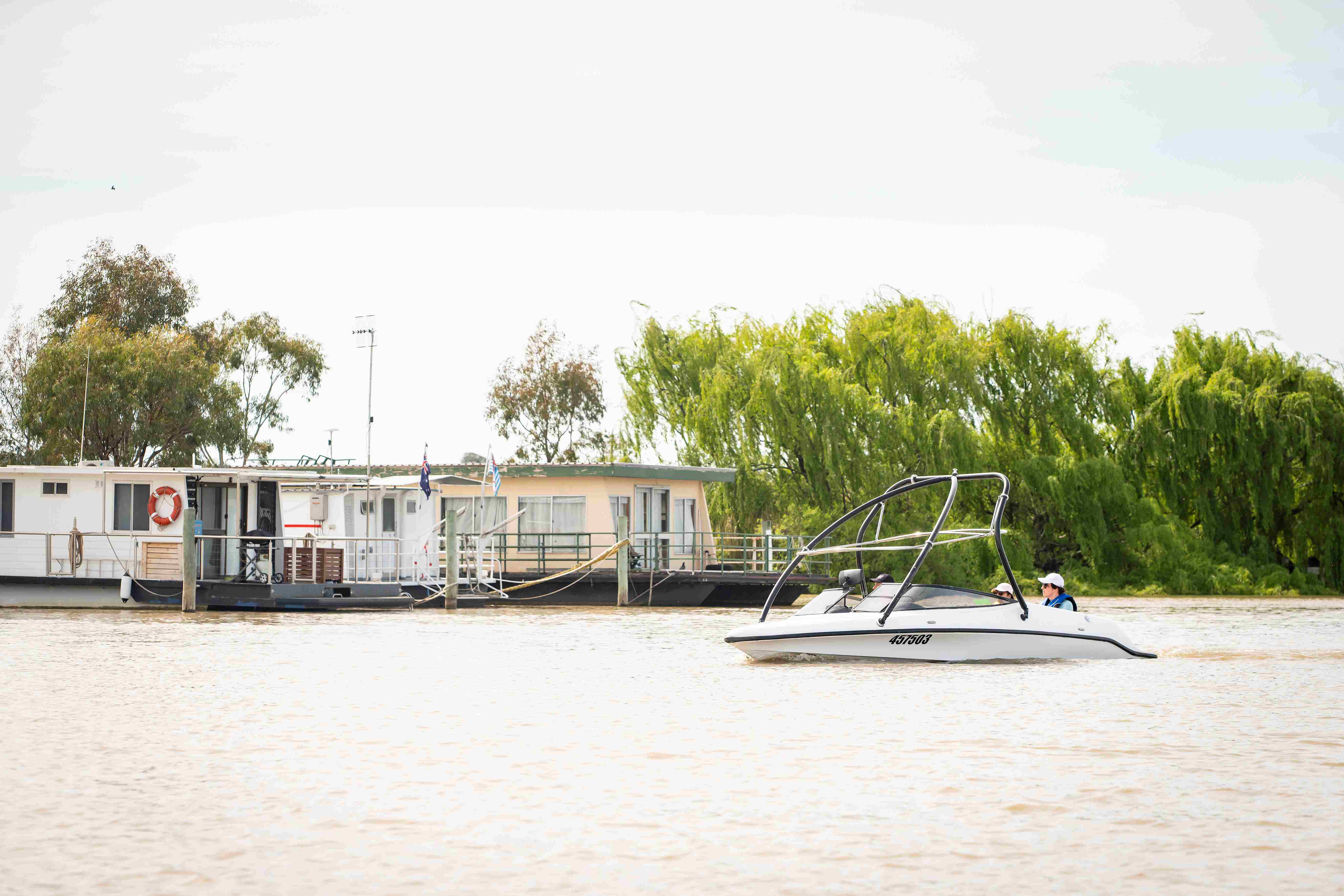 White speed boat traveling on the River Murray with houseboats moored in the background and willow trees next to the houseboats