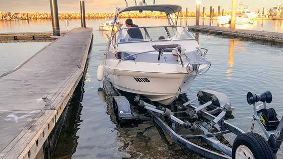 Pictured is a boat and boat trailer half inserted in the water at a boat ramp. Photo has the sun beaming in the background with a few boats in the background in the water