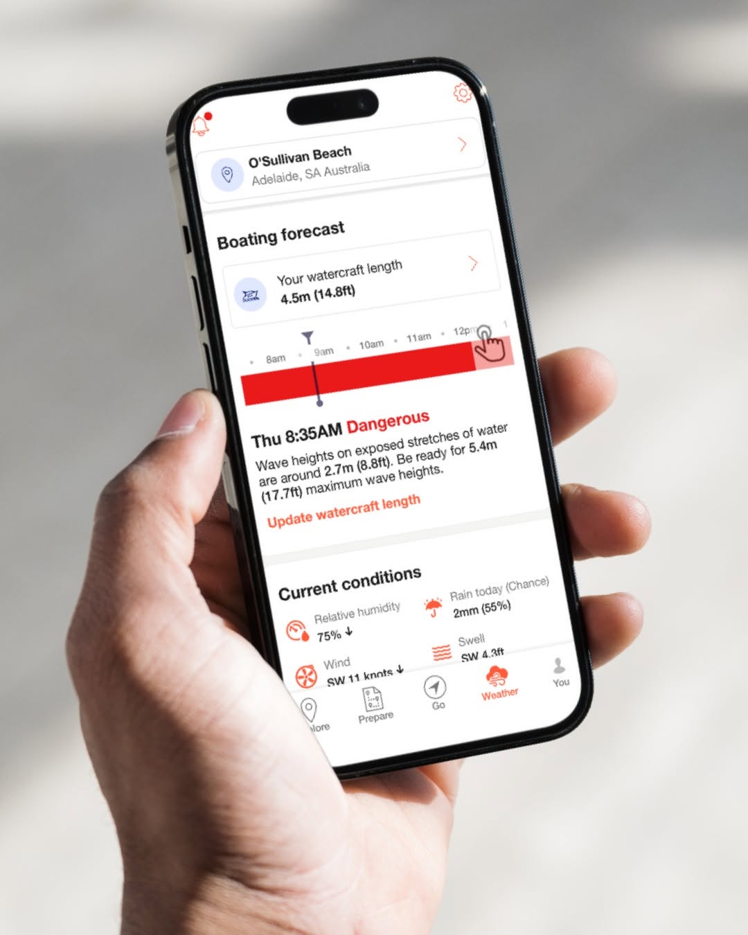 The image shows a smartphone displaying the Deckee boating safety app. The app is open to a location at O'Sullivans Beach, Adelaide, South Australia, with vessel details entered and boating conditions displayed for the day. A prominent warning on the screen indicates that conditions are considered dangerous at a specific time, alerting boaters that waves on exposed stretches of water may exceed the safe operating limits for the selected vessel.  Additional information shown within the app includes wind speed and direction, swell conditions, and other marine weather data to help boaters assess conditions before heading out on the water. The smartphone is held in one hand against a blurred outdoor background, emphasising the convenience of accessing important safety information while on the go. The image highlights how the Deckee app can help recreational boaters make informed decisions and improve safety on the water.
