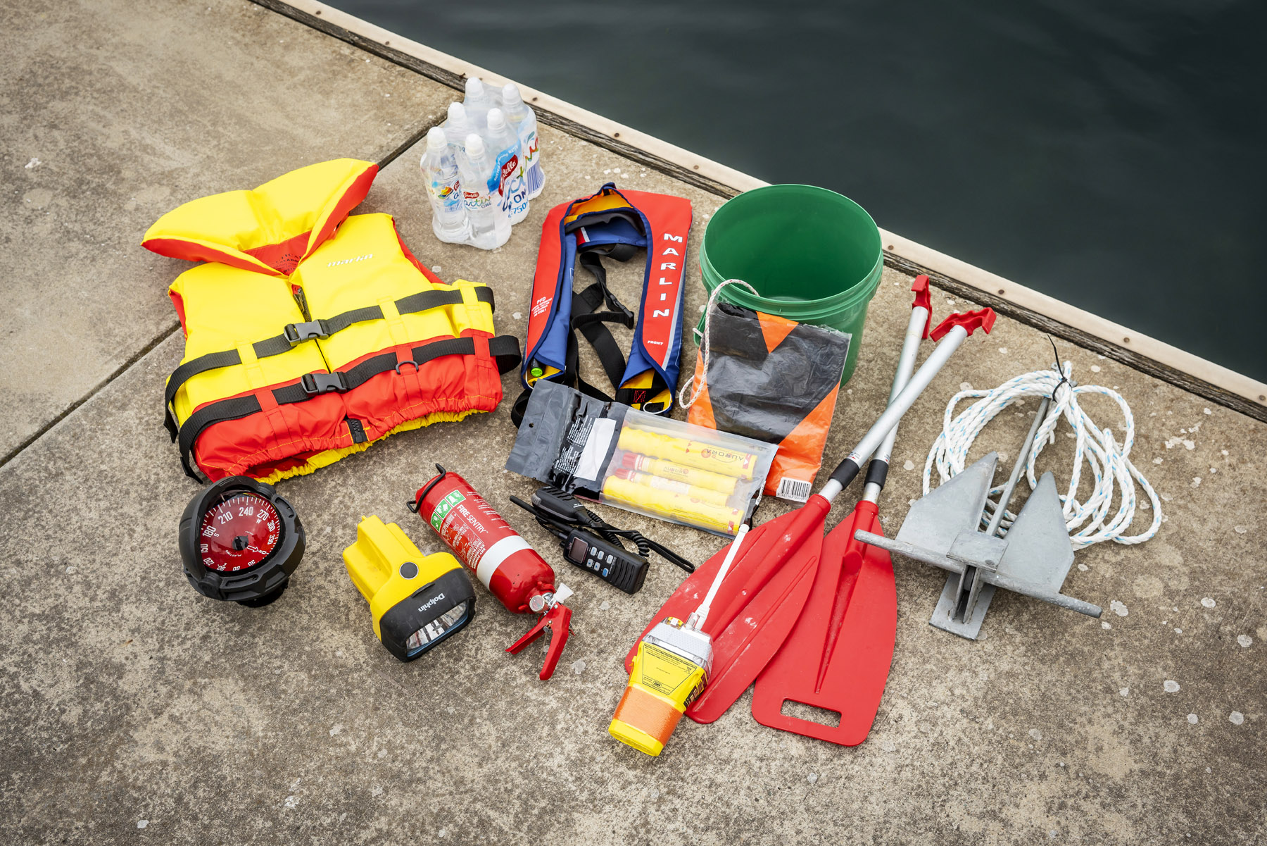 Boating safety equipment laying flat on dock at marina - lifejacket, compass, fire extinguisher, EPIRB, paddles, anchor, bucket, V sheet, flares and radio  