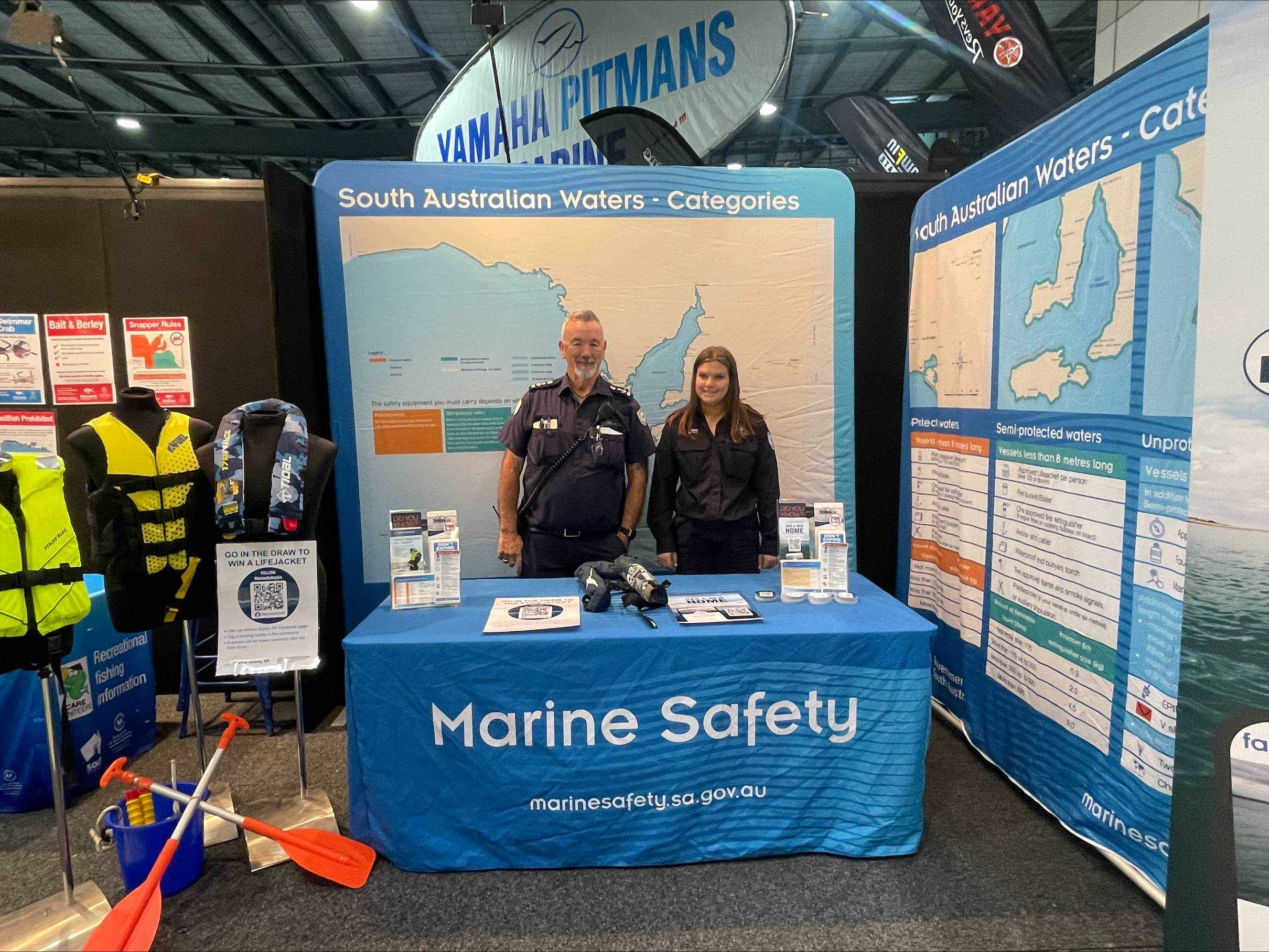 Pictured is two Marine Safety Officers standing at our Marine Safety stand at the 2024 Adelaide 4WD and SA boat & fishing show. To the left is to pole stands with lifejackets and to the right is large posters showing a detailed South Australian Waters Categories.