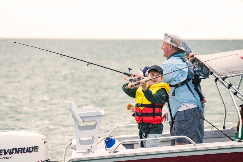 A small recreational boat is floating on calm water. Two people are standing near the side of the boat holding fishing rods. One person is wearing a bright yellow and red lifejacket and appears to be actively reeling in a fishing line, while another person beside them is assisting or fishing alongside. The boat has a canopy overhead for shade and an outboard motor mounted at the rear. The water stretches across the background to the horizon, creating a peaceful coastal boating scene. the weather is over cast with choppy water . The image highlights safe and enjoyable recreational fishing on the water, with lifejacket use clearly visible.