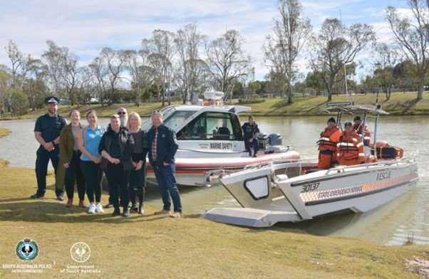 Marine Safety SA team members on one of our boat alongside SES members on their boat. Both boats are on water close to the riverbank, with SA Police and other Riverland stakeholders standing in front of the boats on the bank.