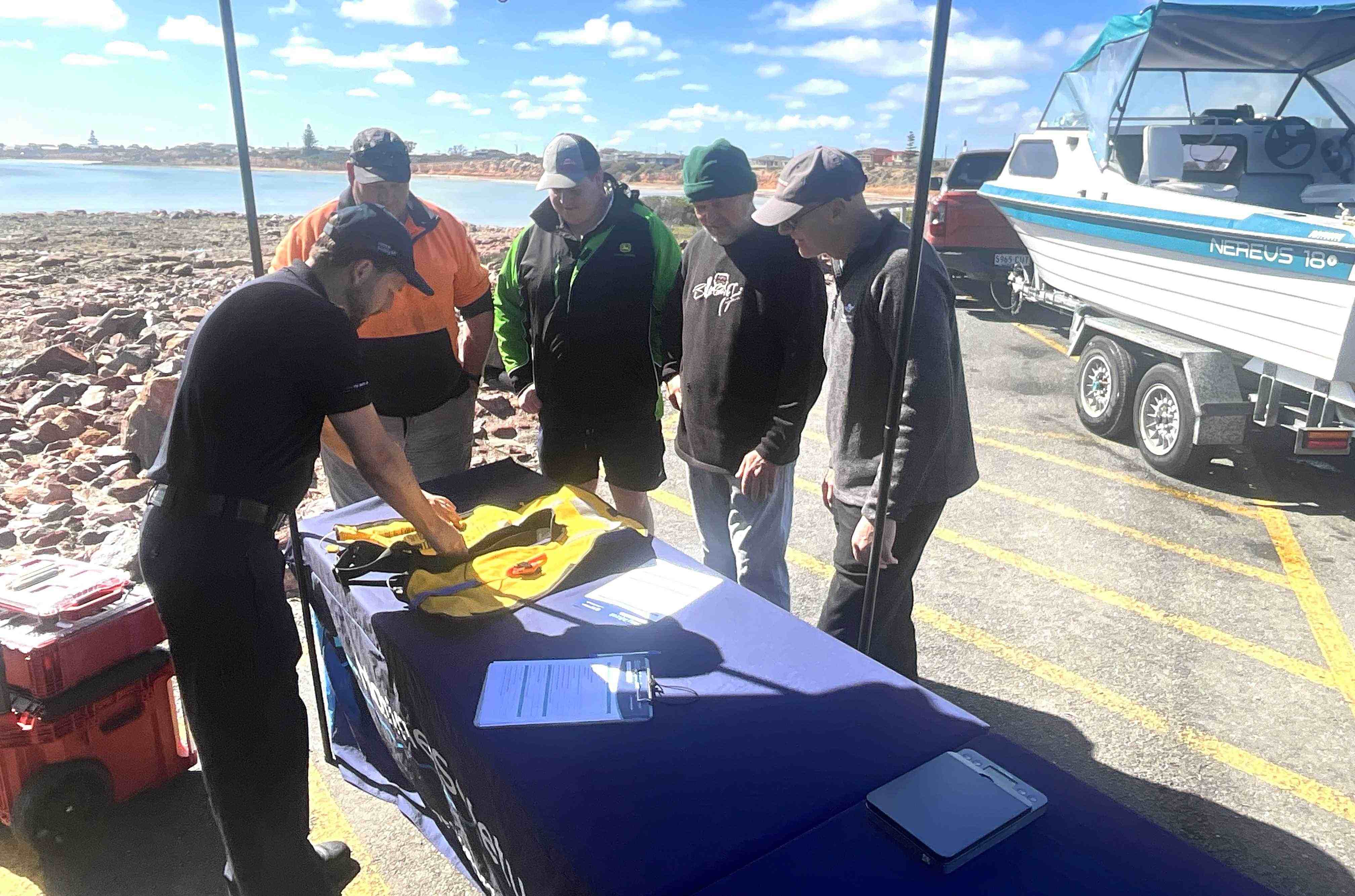 Our Marine Safety Officer showing four gentleman how to maintain / self check your lifejackets. In the background is rocks and ocean.