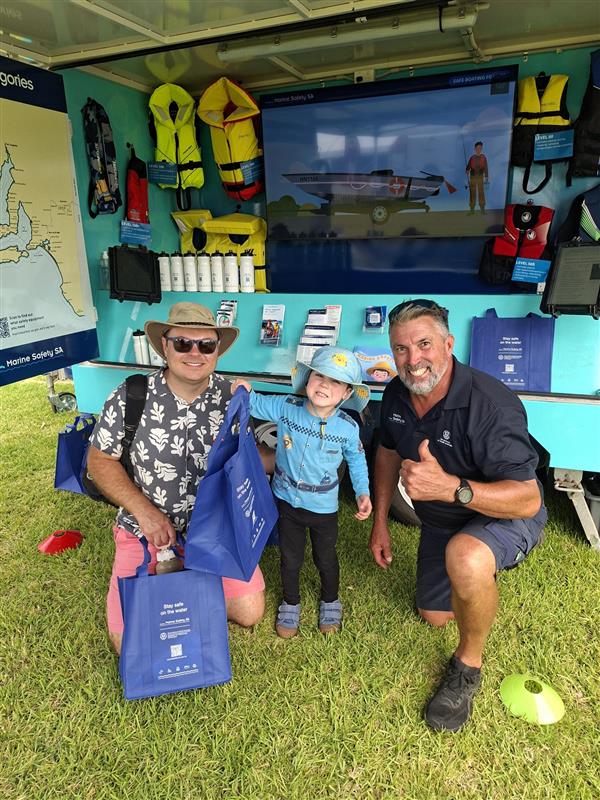 A Marine Safety Officer with a dad and young kid holding a blue Marine Safety tote bag kneeling on grass in front of our Stay Afloat clinic trailer that shows lifejacekts, brochures and drink bottles. It was an overcast day and warm weather.
