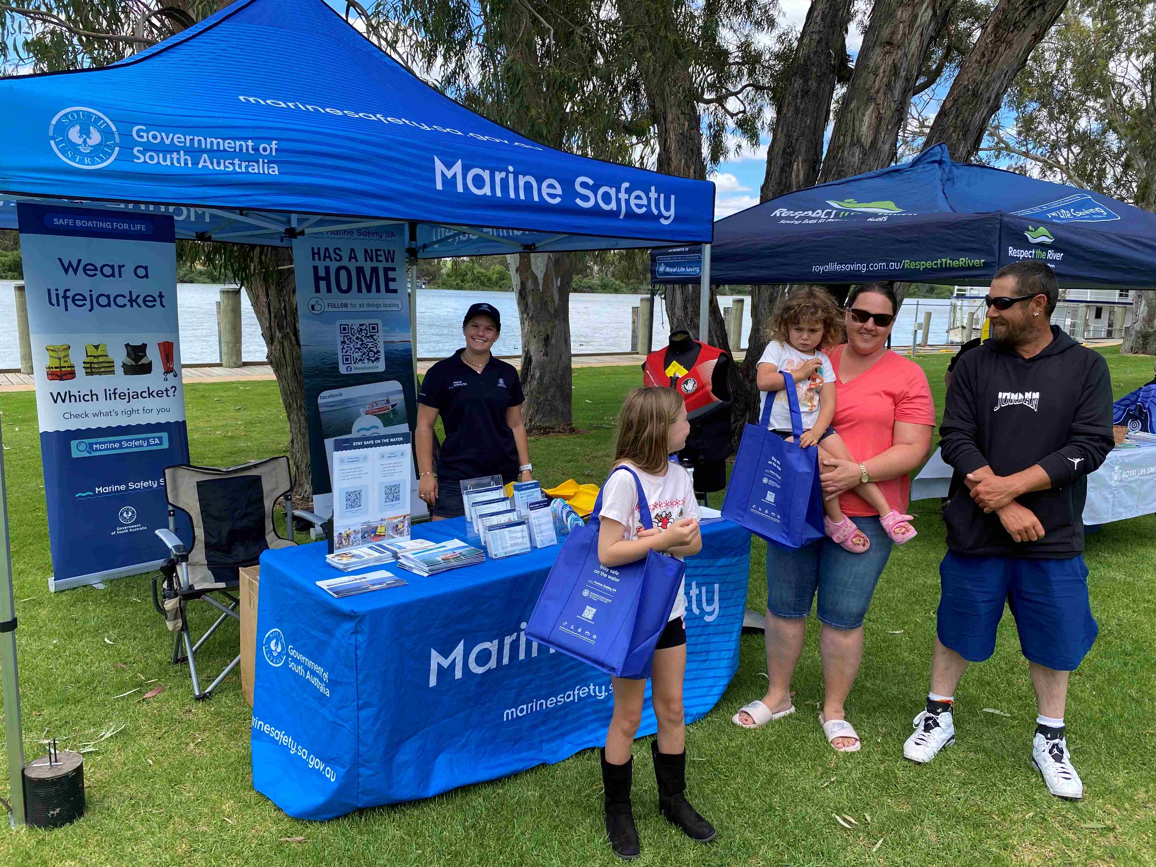 Marine Safety Officer standing with promotional material ona  table with members of the community holding Marine Safety SA tote bags - family of 4 including two adults and two children