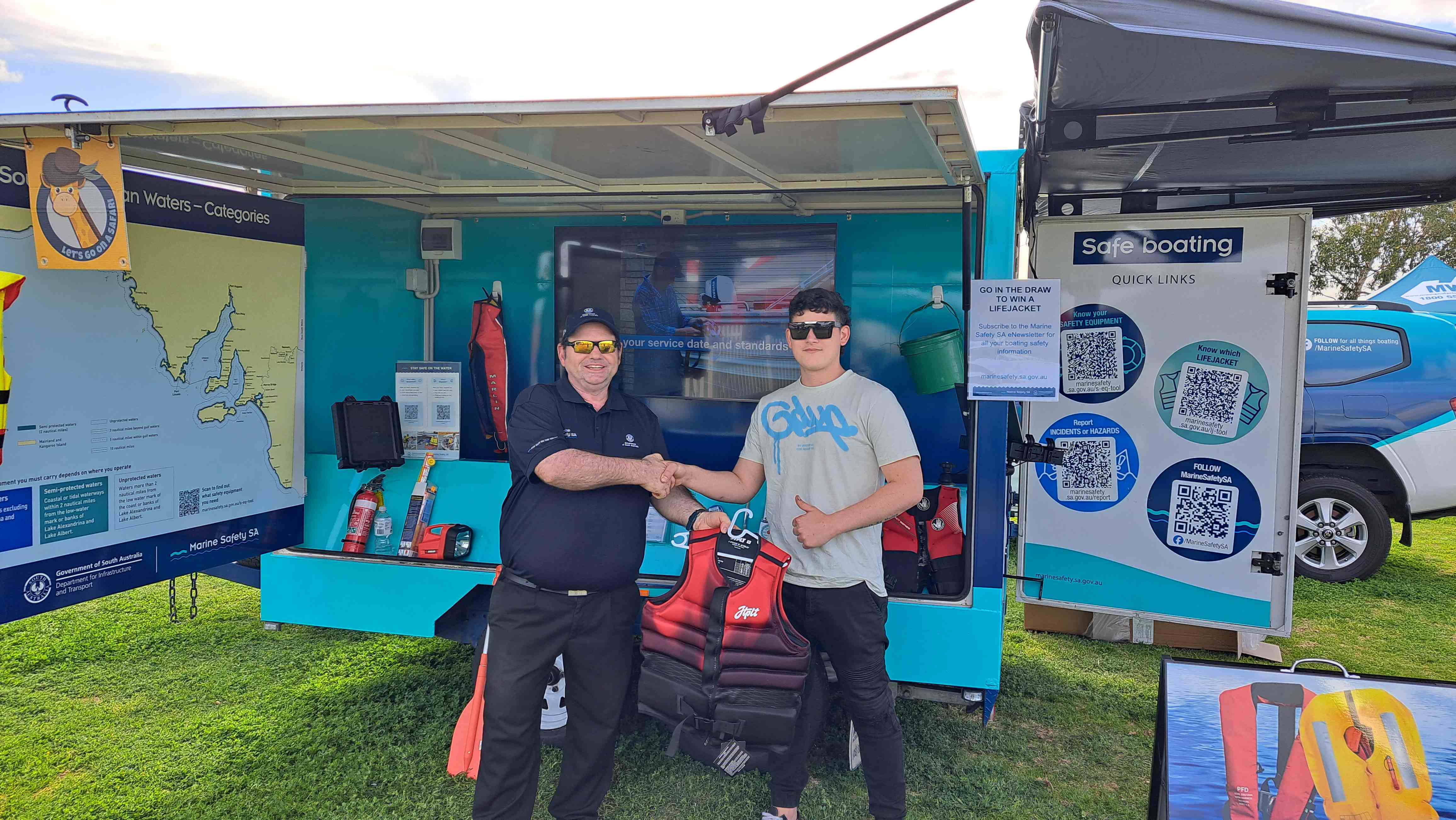 Marine Safety SA staff member presenting man with a new lifejacket and shaking his hand. In the background is the Marine Safety SA education trailer